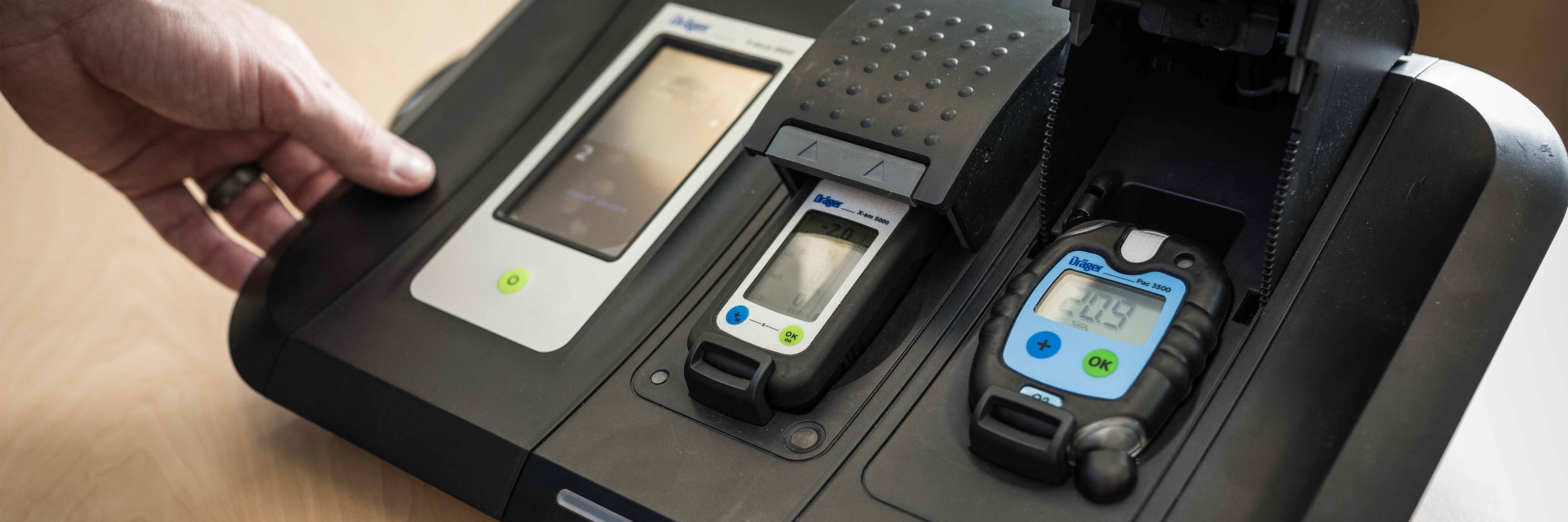 Hand touching a black device tester with two gas detectors on a charging station