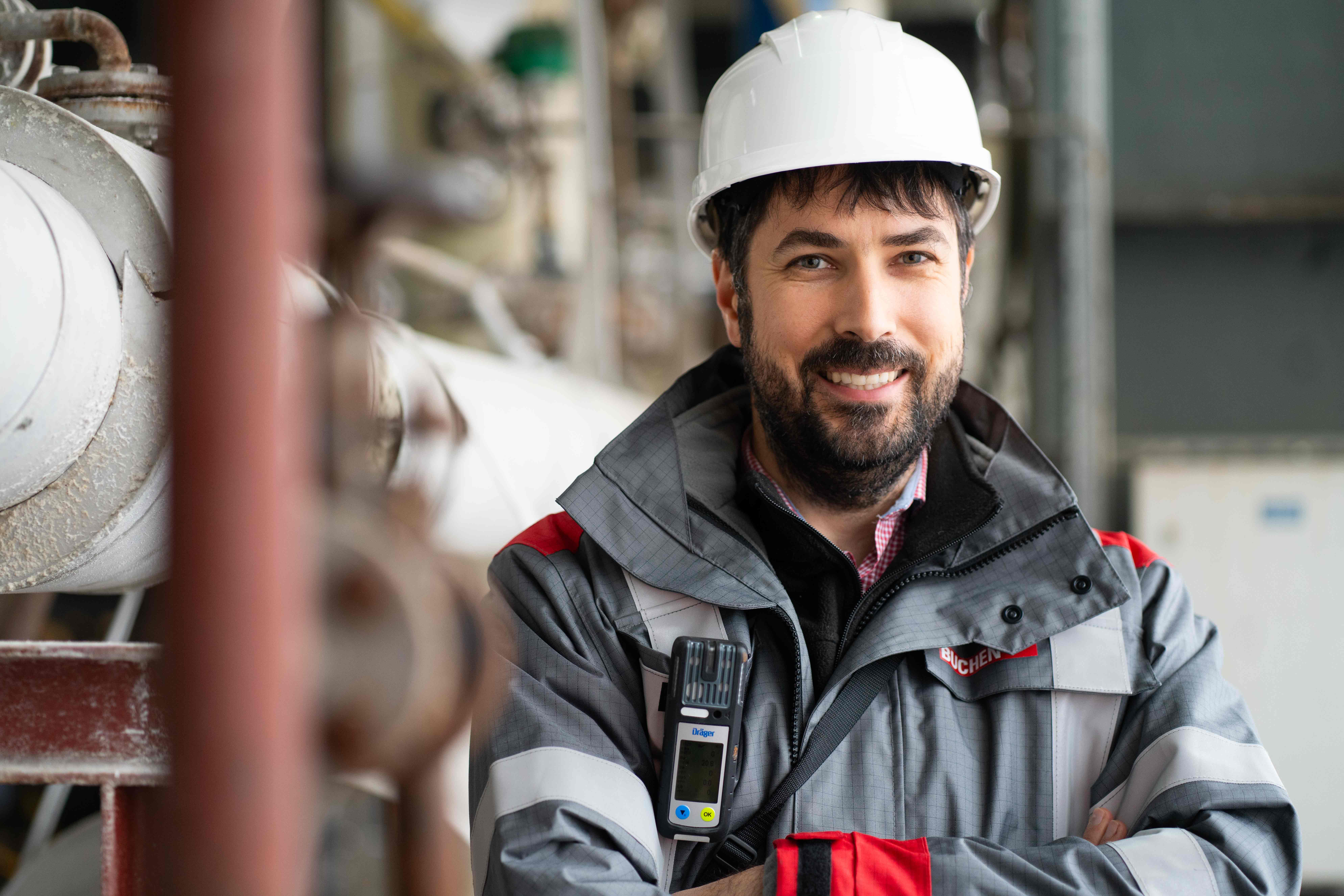 Ein Mann trägt eine Schutzkleidung mit reflektierenden Streifen und einen weißen Schutzhelm. Er hält ein Messgerät an der Brust. Im Hintergrund sind industriell